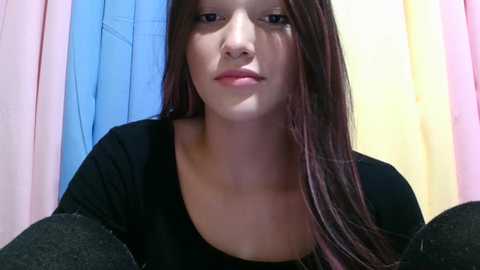 Video of a young, fair-skinned girl with long, dark brown hair, wearing a black top, standing against a backdrop of pastel-colored curtains. Her expression is neutral.