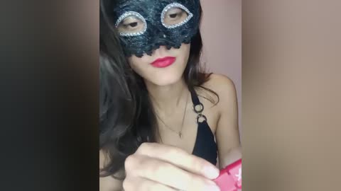 Video of a woman with fair skin, long dark hair, wearing a black lace masquerade mask, bright red lipstick, and a black halter top, with a blurred background.