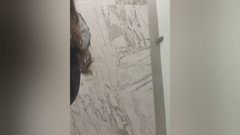 Video of a woman with long, wavy brown hair, partially cropped, standing in a doorway. She wears a blue top and jeans. Marble-tiled walls in the background feature intricate white and gray veins. The image captures a casual, indoor setting.