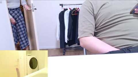 Video: A person in a beige shirt and plaid skirt stands in a doorway, while a person in a beige shirt and black pants sits at a white table with a yellow appliance in the foreground.