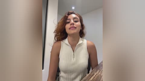 Video of a light-skinned woman with wavy, shoulder-length auburn hair, wearing a sleeveless, ribbed cream top, smiling slightly. Background shows a modern, minimalist room with white walls and a wooden table.