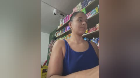 Video of a Latina woman with dark hair in a ponytail, wearing a blue tank top, standing in a colorful convenience store with shelves of snacks and drinks.
