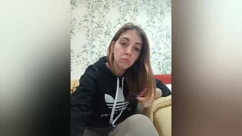 Video of a young woman with straight, brown hair, wearing a black Adidas hoodie, seated indoors with floral wallpaper and a yellow sofa in the background.