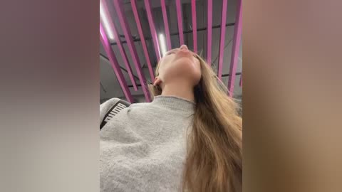 Video of a blonde woman in a gray sweater, leaning back with her head tilted up, surrounded by vertical pink lighting and a modern ceiling.