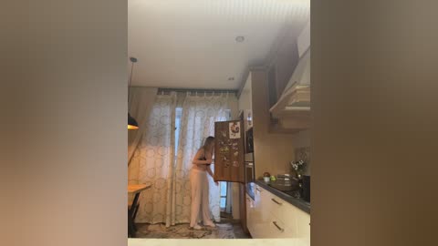 Video of a young woman in a white dress standing in a dimly lit, modern kitchen with beige walls, a stainless steel stove, and a refrigerator.