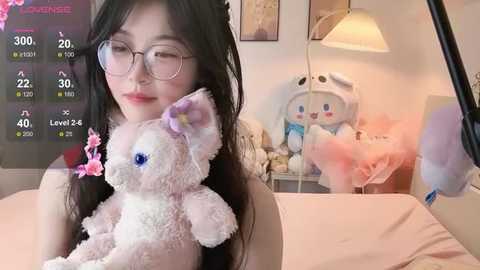 A video of an Asian woman with glasses, long black hair, and a plush pink bunny, in a cozy bedroom with a white lamp, pink wall, and stuffed animals.