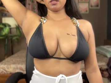 Video of a curvy, light-skinned woman with large breasts, wearing a black bikini top with gold bling and white shorts, posing indoors with plants and framed art in the background.