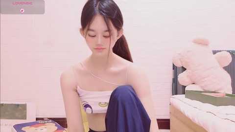 Video of a young Asian woman with light skin, long black hair, and petite frame, wearing a white crop top and blue pants, sitting on a bed with a teddy bear and a green blanket in a pastel-colored room.