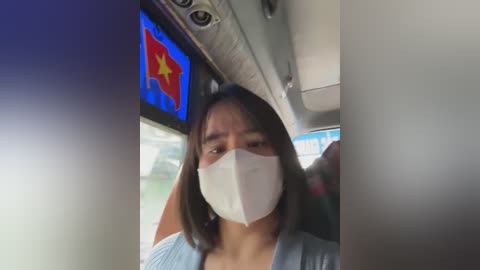 Video of a woman with shoulder-length dark hair, wearing a light blue top and a white face mask, seated in a bus with a blue and red flag hanging above.