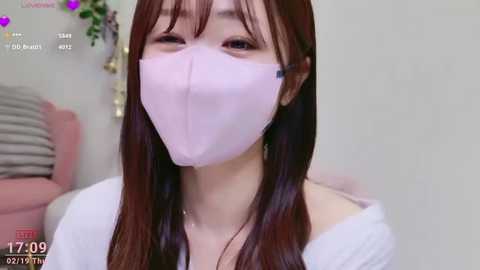 Video of an Asian woman with long, straight brown hair, wearing a light pink face mask and a white off-shoulder top. She has a soft, neutral expression. Background features a pink couch and a green plant.
