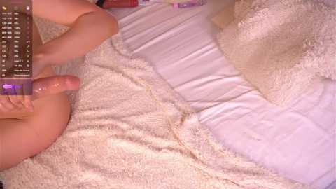 A video of a nude, light-skinned person with a penis, holding a dildo, lying on a bed with white sheets and a fluffy blanket.