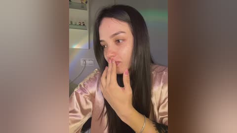 Video of a young woman with long, straight black hair, wearing a shiny pink satin robe, touching her lips with her right hand, set in a dimly lit room with a colorful, blurry background.