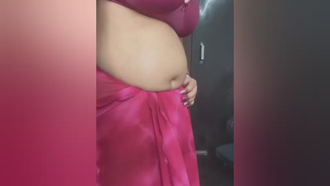 Media: A close-up video of a woman with light brown skin, wearing a pink bra and a matching pink saree, standing in a dimly lit room with dark walls.