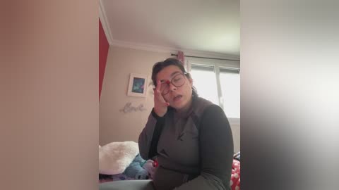 A video of a plus-sized woman with short dark hair, glasses, and a mustache, wearing a black top, talking on a phone, in a cozy, dimly lit room with a red wall and a window.