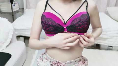 Video of a slender, light-skinned woman in a pink and black lace bra, adjusting the straps, standing in a tidy bedroom with white furniture and floral-patterned pajama pants.