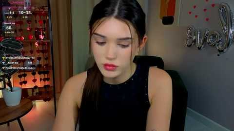 Video of a young woman with straight dark hair, wearing a black sleeveless top, seated in a dimly lit room with a wooden table and a string of red hearts.