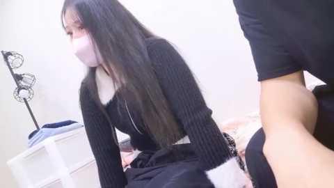 A video of an Asian woman with long black hair, wearing a face mask, sitting on a bed with a man in black clothes, in a white room with minimalistic decor.