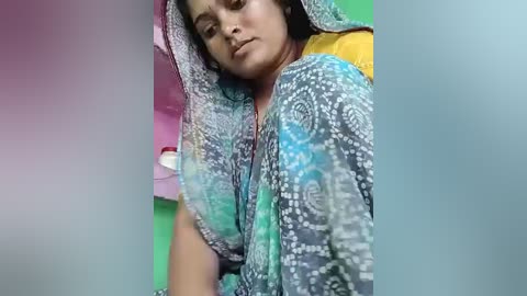 Media: A video of a South Asian woman with medium brown skin, wearing a colorful, patterned saree, lying on a bed with a green and yellow backdrop.