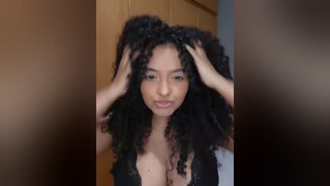 Video of a young woman with a medium complexion, large breasts, and voluminous curly black hair, wearing a black lace bra. She is indoors with wooden paneling in the background, hands in her hair.