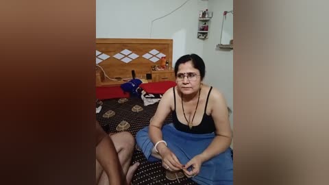 Video of a woman with glasses and dark hair, wearing a black tank top and blue pants, sitting on a bed with a patterned blanket. Background includes a wooden headboard with a white geometric design, a red bedspread, and a cluttered bedside table.