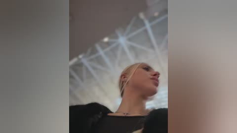 Video of a fair-skinned woman with blonde hair, wearing a black top, standing in a modern indoor space with a white, geometric ceiling and large windows.