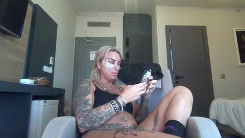 A tattooed, blonde woman in a black bra and panties, sitting on a white chair, plays with a smartphone in a modern, minimalist room.