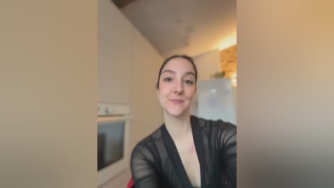 A video of a young woman with fair skin and dark hair, wearing a sheer black robe, standing in a modern, beige-tiled kitchen with stainless steel appliances and a stone backsplash.