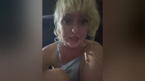 A close-up video of a young woman with platinum blonde, messy hair, wearing a light blue tank top, looking directly at the camera with a neutral expression, slightly out of focus.