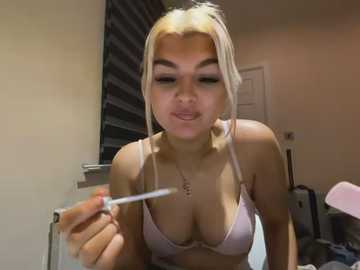 Video of a young woman with platinum blonde hair, wearing a pink bra, applying lip gloss in a bedroom.