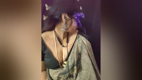 A video of a woman with a medium-dark complexion, wearing a sheer, green paisley-patterned dress with a deep neckline, adorned with multiple necklaces, including a long pearl strand. Her face is partially obscured by a dark, patterned veil. The background is a blurred, dark purple.