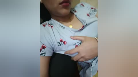 A close-up video of a person with light skin, wearing a white shirt with cherry prints, and a necklace, holding their chest. The background is blurred.