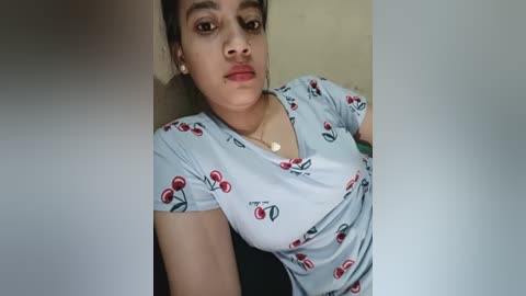 Video of a young South Asian woman with light brown skin, dark hair in a ponytail, wearing a light blue t-shirt with cherry print, indoors, neutral background.