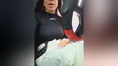 A video shows a woman in a black sports bra and beige pants, with a visible large stomach, seated in a car, blurred background.