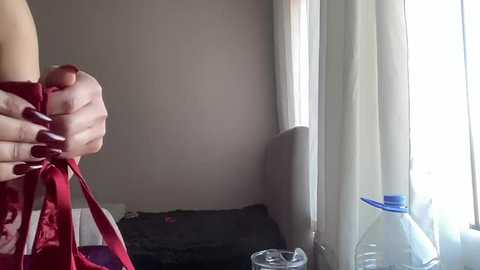 Video of a person's hand holding a red ribbon, standing near a window with white curtains, partially visible. The room is dimly lit with a dark gray couch and a blue water bottle in the foreground.