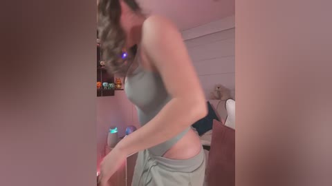 Video of a woman with wavy brown hair, wearing a grey tank top and grey shorts, adjusting her clothes in a dimly lit room with a shelf of books and a desk.