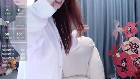 A video of an Asian woman with straight, shoulder-length red hair, wearing a white blouse, standing in front of a backdrop featuring red and black floral patterns.