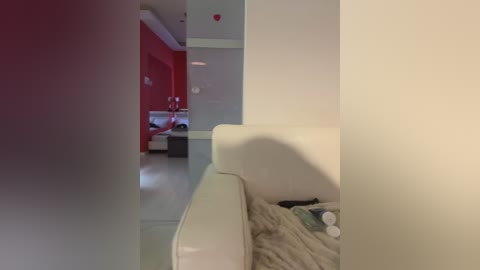 Video of a modern, minimalist living room with white leather couch and red accent wall, leading to a dimly lit bedroom with a bed.