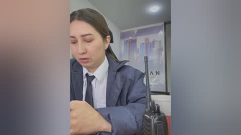 Video of a young Asian woman in a dark navy coat, white shirt, and black tie, looking serious, speaking into a handheld radio, in a modern office with a poster of a city skyline in the background.