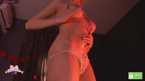 A video of a topless, light-skinned woman with medium-sized breasts, wearing only white lace panties, standing in a dimly lit room with red lighting.