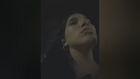Video of a woman with dark hair and closed eyes, wearing gold hoop earrings, in a dimly lit room with dark and light brown tones.