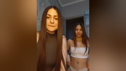Video of two young women with long, straight brown hair, one in a sheer black top, the other in a white crop top, standing side by side in a bathroom with a tiled wall.