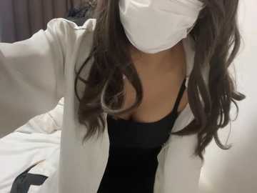A close-up video of a woman with long, wavy brown hair, wearing a white mask, black top, and white jacket, taking a selfie. Background shows a bed with white sheets and a blurred dark object.