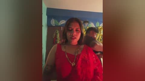 Video of a South Asian woman with medium skin tone, wearing a red saree, standing indoors, with a colorful wall mural in the background.
