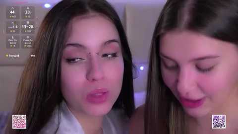 Video of two young women with fair skin, dark hair, and makeup, kissing in a dimly lit room with a hospital sign.