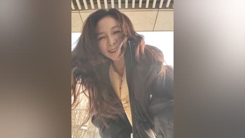 Video of a young Asian woman with long, wavy brown hair, wearing a dark jacket, laughing with her head tilted back, in a modern indoor setting with light wooden paneling and a beige ceiling.