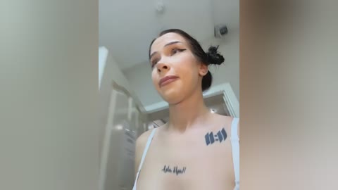 Video of a young woman with fair skin, dark hair in a bun, wearing a white spaghetti-strap top. She has tattoos on her shoulders and chest, standing in a modern, white-walled bathroom with a mirror and shower.