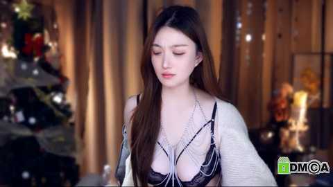 A video of a young woman with long brown hair wearing a sheer, white lace bra with black detailing, standing in a softly lit, festive indoor setting with Christmas decorations.