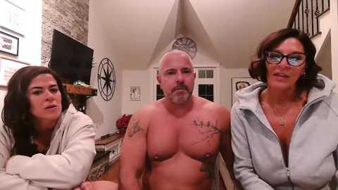 Video of three people in a cozy, modern living room: a bald, muscular man with tattoos, a woman with glasses, and another woman with long hair.