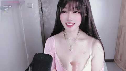 Video of a smiling Asian woman with long black hair, wearing a pink top, holding a black microphone, in a dimly lit room.