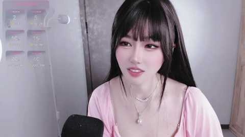 A video of an East Asian woman with long black hair, wearing a pink blouse, speaking into a microphone in a studio setting.
